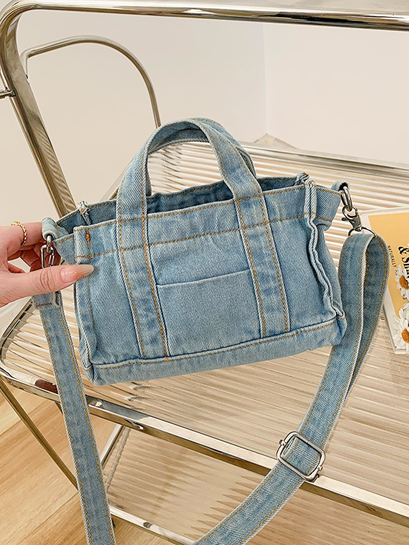 denim shoulder handbag, light blue, held at an angle by a woman's hand, on a glass and metal slatted cart